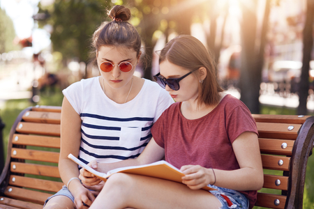 Two best friends or classmates prepare for final exams at university wears trendy sunglasses t shirt have look into book sit at bench in park enjoy sunny weather during spring teen girls studying