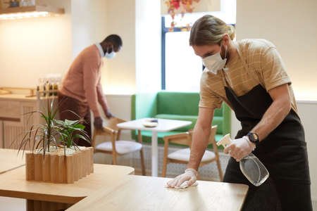 Retrato de un trabajador de café masculino con máscara y protector facial mientras desinfecta mesas y limpia muebles, copia espacio