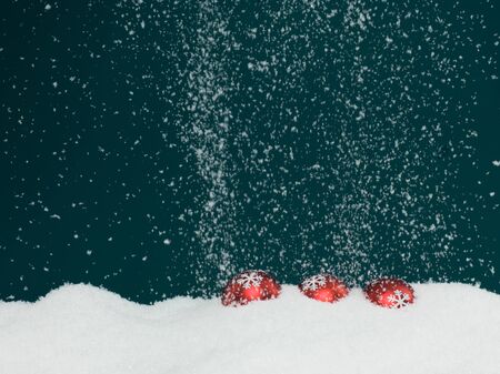 Closeup of three red colored christmas globes decorated with glittery snowflakes surrounded with snow on blue background with falling snowflakes