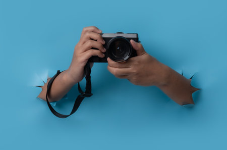 Female hand holding camera breaks through blue paper background