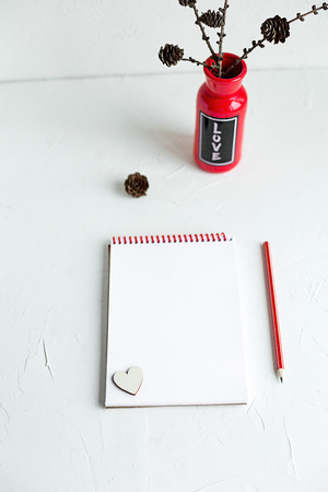 An open notebook with red pencil and a red vase with love on it are on white table st valentine s day concept