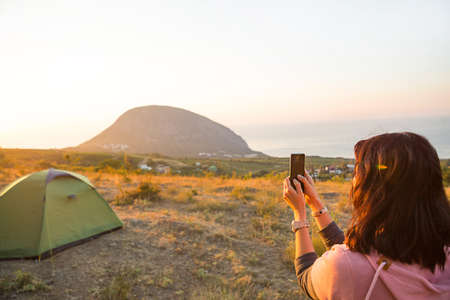 Mujer toma fotos del amanecer en las montañas en su teléfono. Selfie en sol naciente. Vista panorámica sobre el mar y Ayu-Dag. Camping, actividades al aire libre, senderismo deportivo de montaña, viajes en familia. Crimea.