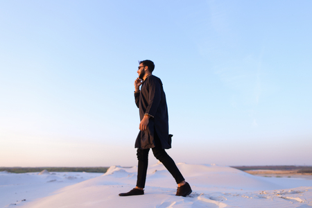 Handsome muslim businessman conducts dialogue on smartphone with business partner and tells ideas for progressing matters or solves important work issues while standing in middle of bottomless desert with white sand on warm summer evening swarthy muslim Stock Photo