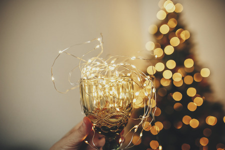 Hand holding stylish vintage glass with garland bulbs inside on background of golden beautiful christmas tree with lights in festive room cozy winter holidays cheers for christmas Stock Photo