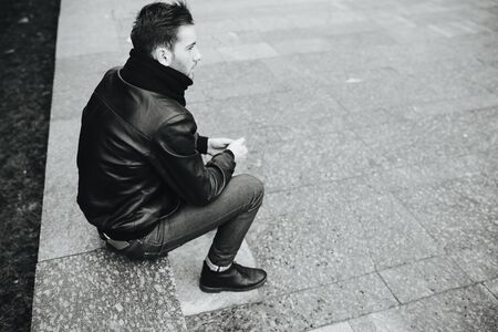 A man dressed in jeans and black jacket seats on a slab