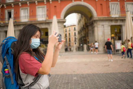 nuevos viajes de vacaciones normales en Europa - joven hermosa y feliz turista china asiática con máscara facial y mochila tomando fotos con teléfono móvil disfrutando de un recorrido por la ciudad emocionada