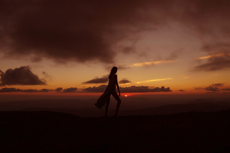 Pretty girl slim model silhouette with hair poses in summer dress on mountain top on beautiful sunset sky natural background Stock Photo