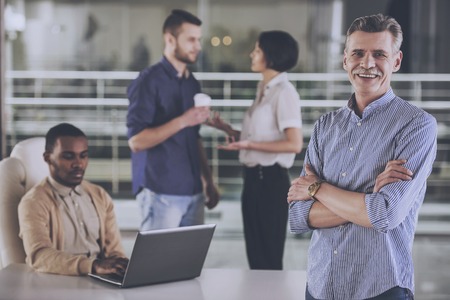 El jefe sonriente y su equipo están trabajando en la oficina. Jefe con las manos cruzadas. Gente de negocios en segundo plano. Proceso de trabajo. Hombre de pie. Señalamiento corporativo. Jefe con colega. Grupo profesional.