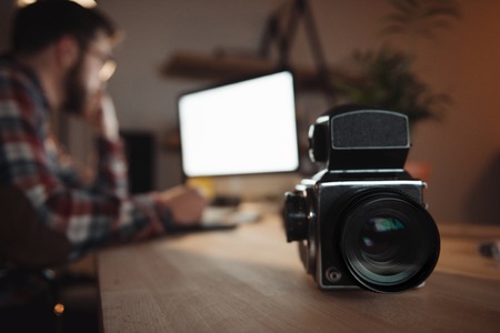 Photo of camera standing on table near bearded web designer dressed in shirt in a cage print and wearing glasses focus on camera