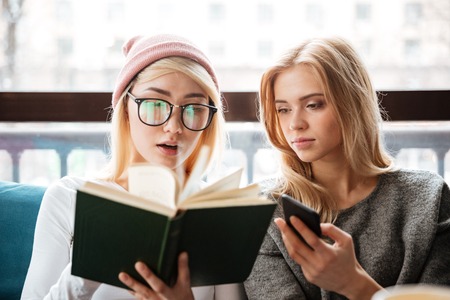 Image of young cheerful two women friends sitting in cafe and reading book while holding phone Stock Photo