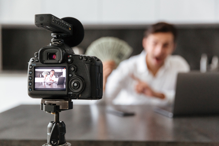 Happy young man filming his video blog episode about new tech devices while sitting at the kitchen table with laptop and showing bunch of money banknotes