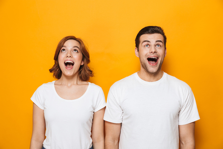 Hermosa pareja emocionada con camisetas blancas aisladas sobre fondo amarillo, mirando hacia arriba