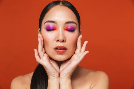 Beauty portrait of an attractive young asian woman with brunette hair wearing bright makeup standing isolated over red background eyes closed posing Stock Photo