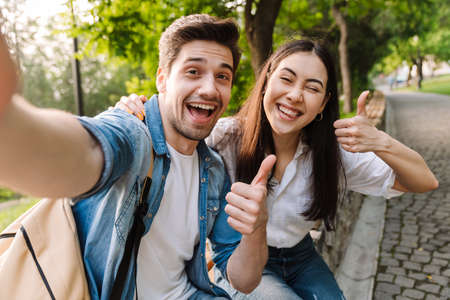 Image of joyful multicultural couple taking selfie photo and showing thumbs while sitting on bench in park