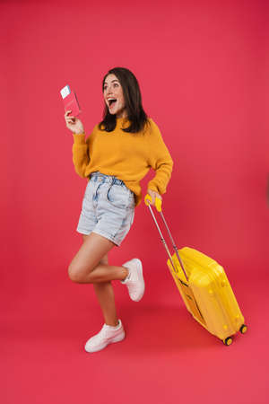 Longitud total de una mujer joven feliz caminando con la bolsa de viaje, con pasaporte y boletos aislados sobre fondo rosa