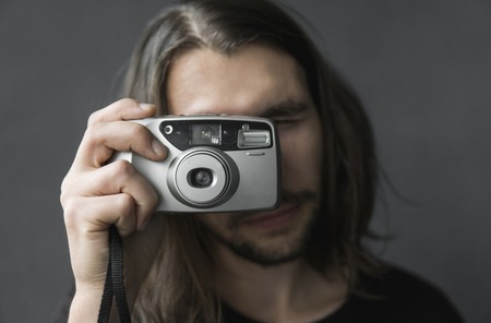 Handsome young bearded man with a long hair and in a black shirt holding vintage old fashioned film camera on a black background and looking in camera viewfinder