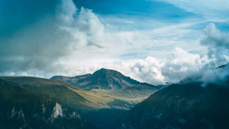 A mountain gorge covered with a forest a mountain with a glacier rises in the background white clouds cling to the mountains blue sky in the background the beauty of nature Фото со стока