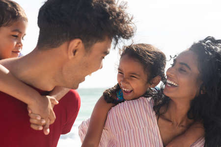 Alegres padres multirraciales dando paseos a cuestas a los niños en la playa en un día soleado. familia, estilo de vida y fin de semana. Foto de archivo