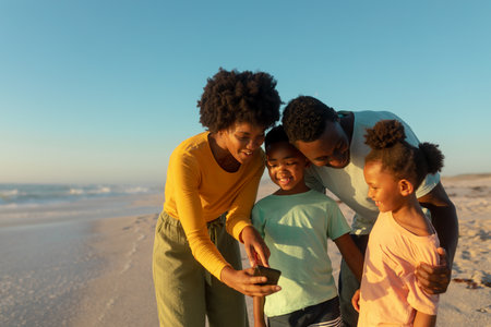 Feliz madre afroamericana compartiendo teléfono inteligente con la familia en la playa en un día soleado. inalterado, familia, estilo de vida, unión, tecnología y concepto de vacaciones. Foto de archivo