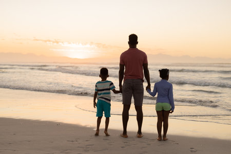 Vista trasera de un padre afroamericano con un hijo y una hija mirando el mar contra el cielo durante la puesta de sol, la naturaleza, la playa inalterada, la infancia, la unión familiar, el estilo de vida, el disfrute y las vacaciones Foto de archivo