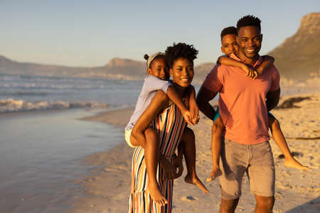 Portrait of happy african american young parents piggybacking son and daughter at beach against sky nature copy space unaltered childhood family togetherness lifestyle enjoyment holiday