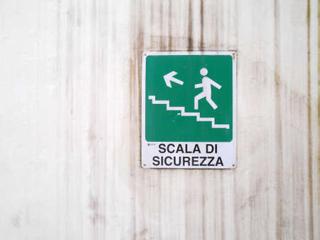 Señal de salida de emergencia Stairs Left Up en italiano en una pared blanca con rayas de tierra. Lagos en Roma Foto de archivo