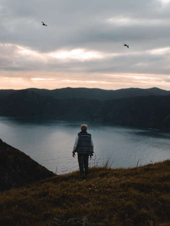 A vertical shot of a person standing on grass by the water near mountains under a pinkish sky with birds flying