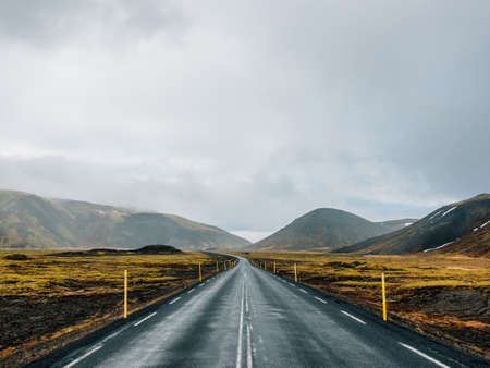 A road surrounded by hills covered in greenery and snow under a cloudy sky in iceland