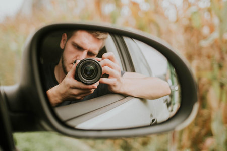 A male taking a selfie in the rearview mirror of the car with a photo camera
