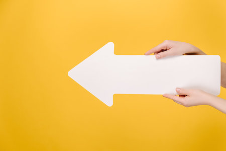 Close up side view of beautiful female hands holding big white paper arrow isolated on yellow studio background with copy space for advertisement advertising area mockup Stock Photo