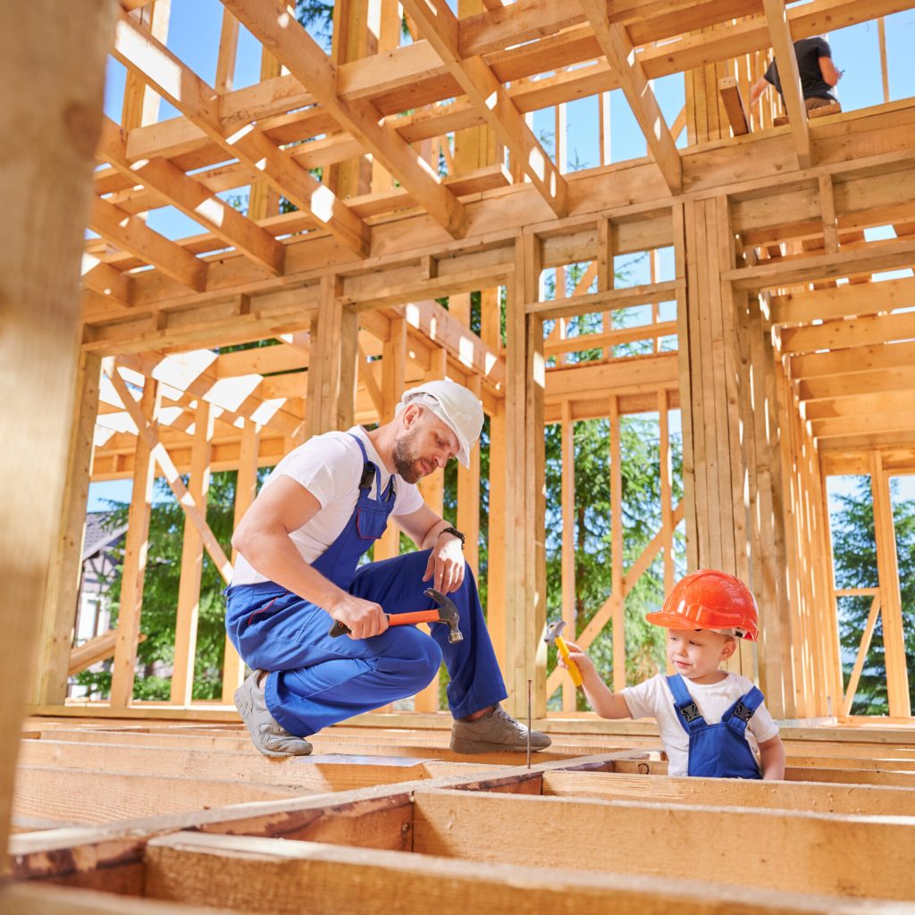 Father with toddler son constructing wooden frame house. Man builder instructing his son on art of hammering nails on construction site, wearing helmets and blue overalls. Carpentry and family concept