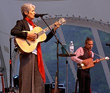 Baez plays outdoors in brown wide-leg pants, white top, brown waistcoat, blue pearls, and a long orange neck scarf. To her left, a male accompanist in a vest plays a small wooden cigar-box-style guitar