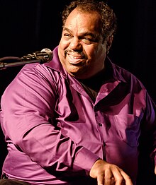 Daryl Davis wearing a purple button-down shirt, playing a piano, turned right-of-camera, towards a microphone