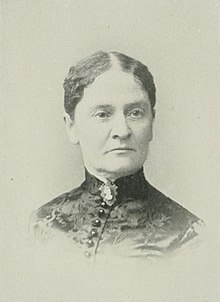 B&W portrait photo of a woman with her hair parted in the center and in an updo, wearing a dark, high-collared blouse.