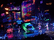 Bird's eye view of a stage, showing large scaffoldings, neon signs and a green car lying in the middle.