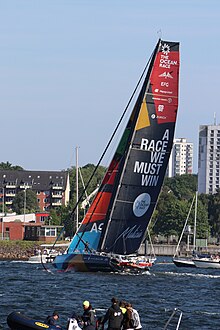 Bateau progressant à la voile, vu de trois quarts arrière, non loin d'une rive urbanisée.