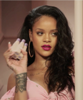 Close-up image of a black woman holding make-up bottles while looking to her left. She wears a pink shirt and has wavy black hair.