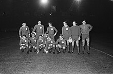 Black and white photo of a football team in kit, with a black sky in the background. Five players are crouched down, and six are standing.
