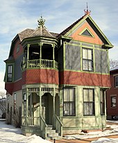 Two-story green home with some orange-red sections in Victorian style with dramatic rooftop ornaments, open second floor turret porch