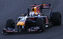 A grey-and-red single-seater racing car is driven around a tarmac course while the driver sticks one of their arms in the air in celebration.