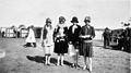 A black and white photo of four women standing side by side in a field. They all wear loosely fitting shirts and knee-length skirts.