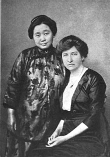 A portrait of two women. The Chinese woman, Shi Meiyu, is standing and wearing a printed silk top; the white woman, Jennie V. Hughes, is seated with her hands in her lap; she is wearing a black dress with a white front. Both women have their hair dressed up off their necks.