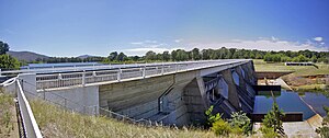 The dam is surrounded by grass and a road runs on top of it. A forest is in the background and there is no human activity in the range of visibility.