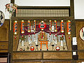 A large, decorative alcove shrine, with red paper lanterns decorated with black text strung across the top, a miniature torii gate in front and two red daruma dolls at either side.