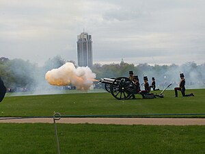 A cannon of the King's Troop Royal Horse Artillery firing in Hyde Park. A large plume of orange smoke, lit orange by the flash from the gun, extends from the barrel.