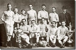 A group of twelve men, eleven in association football attire typical of the late nineteenth century and one in a suit and bowler hat. They are displaying a number of trophies.