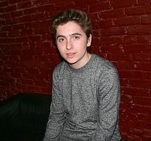 close-up of Gavin Becker wearing a gray t-shirt, sitting and grinning directly at camera, with red-brick wall in background