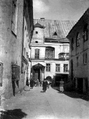 Frontale Schwarzweißfotografie einer Gasse mit einem dreistöckigen Gebäude im Hintergrund. Am Dach ist ein kleiner Turm und im Obergeschoss ist ein großes Fenster mit Holzgitter. Der Eingang besteht aus einer Doppeltür mit einem Giebelfeld. Vor dem Gebäude laufen einige Menschen.