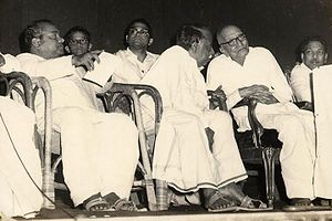 five men and a boy sitting in chairs. Four of the men are middle aged and one is in his seventies. One of the middle aged men is leaning toward and speaking to the old man.