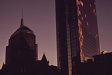 Two dark buildings rise into the early evening sky. The tower on the right is spotted with plywood on its side.